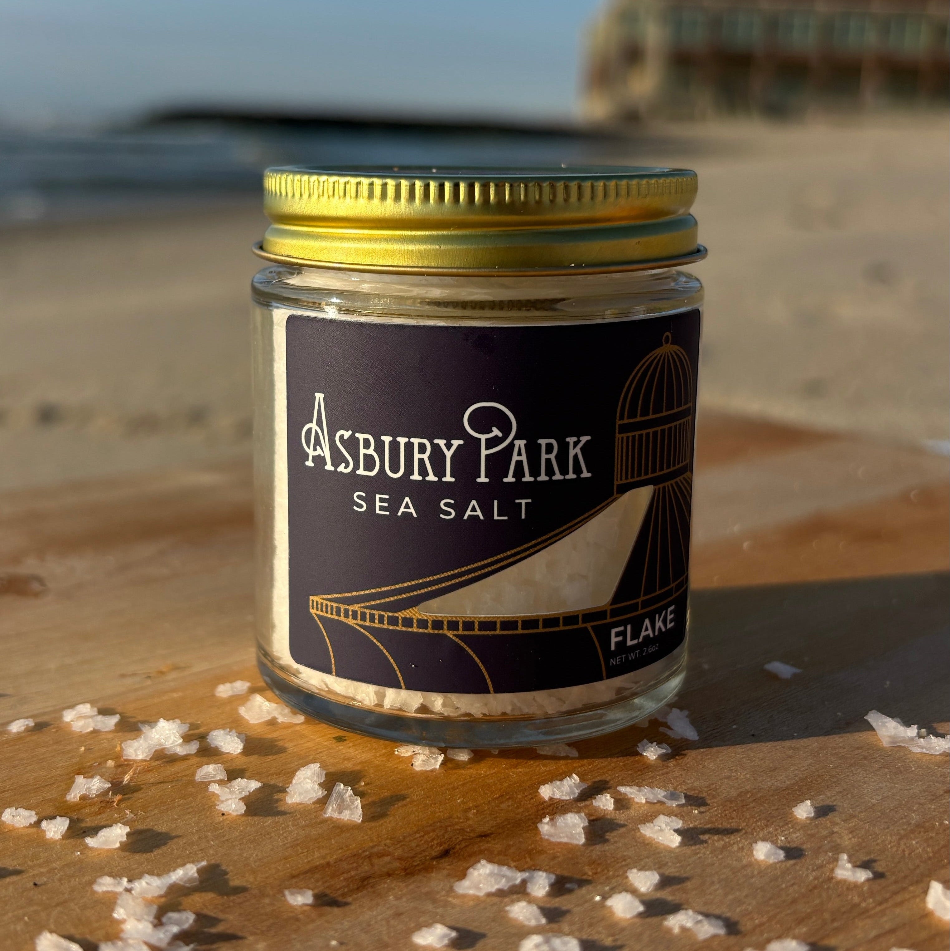 A small glass jar of Asbury Park Sea Salt with a gold lid and navy blue label featuring an image of the sits on a wooden surface outdoors on a beach, with coarse salt crystals scattered around it and a blurred building and blue sky in the background.