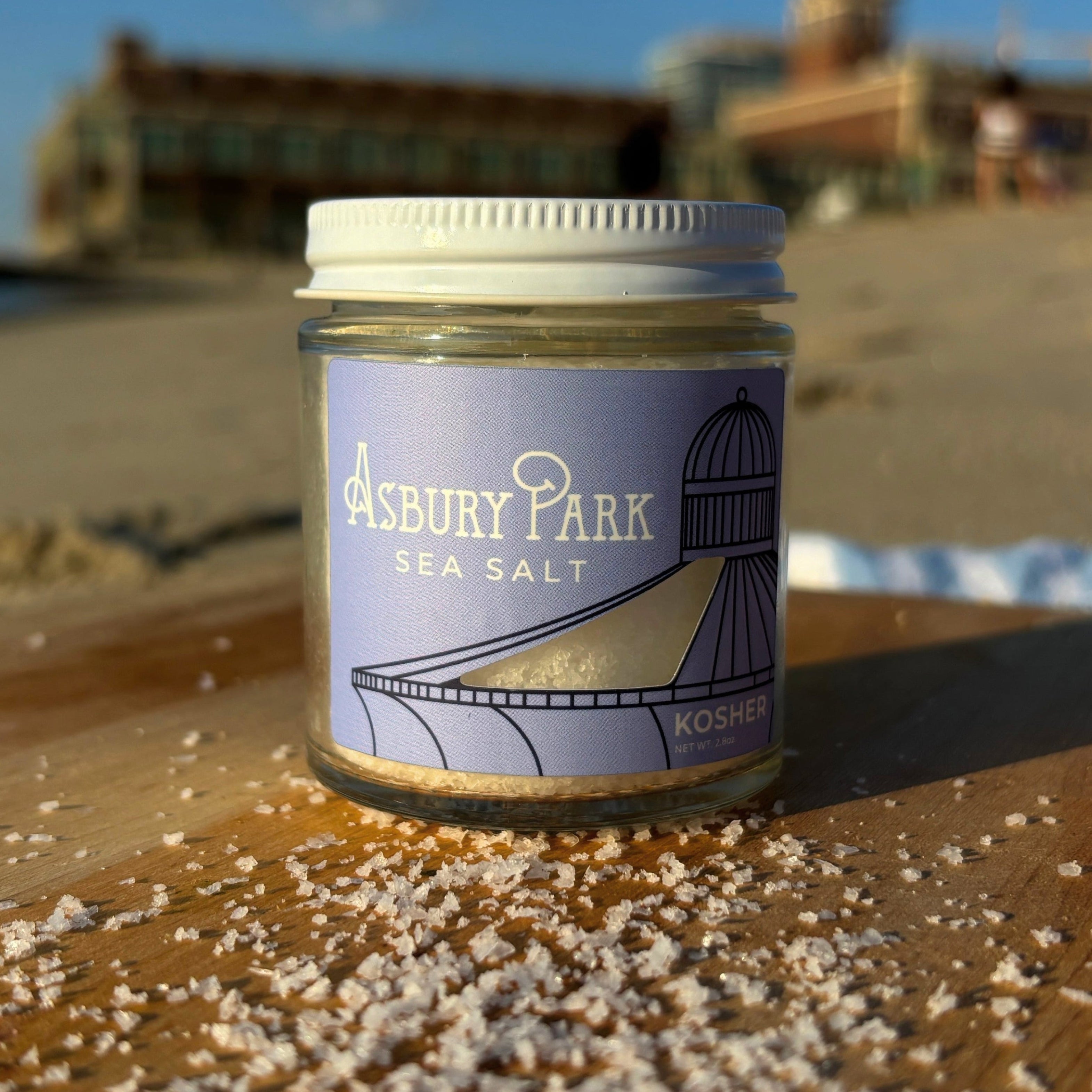 A jar of Asbury Park Kosher Sea Salt sits on a wooden cutting board on a beach, with kosher salt scattered around the base and a blurred background of sand, water, and Convention Hall under a blue sky.