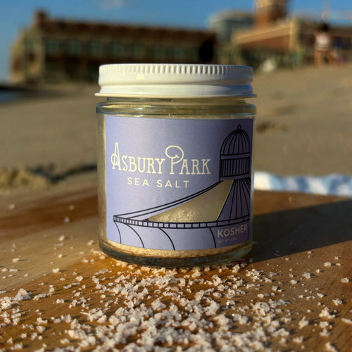 A jar of Asbury Park Kosher Sea Salt sits on a wooden cutting board on a beach, with kosher salt scattered around the base and a blurred background of sand, water, and Convention Hall under a blue sky.